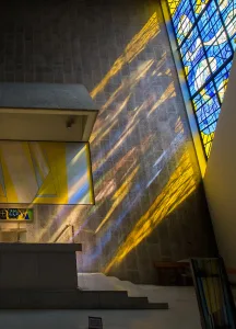 Diagonal bands of yellow and blue light from a tall stained-glass window fall across a textured stone wall above a simple altar and stepped platform in a modern chapel.