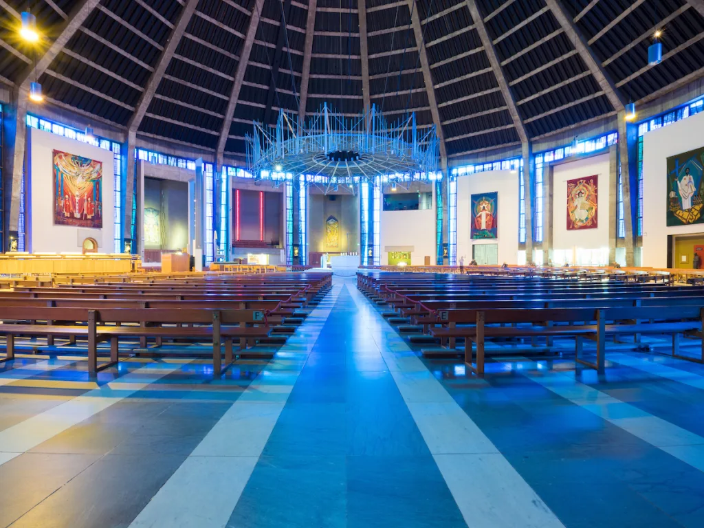 Interior of a large circular modern church with a high ribbed dome, rows of wooden pews flanking a long central aisle leading to a raised altar beneath a suspended spiky metal chandelier, blue-tinted light from tall stained-glass windows and colourful religious murals on the walls.