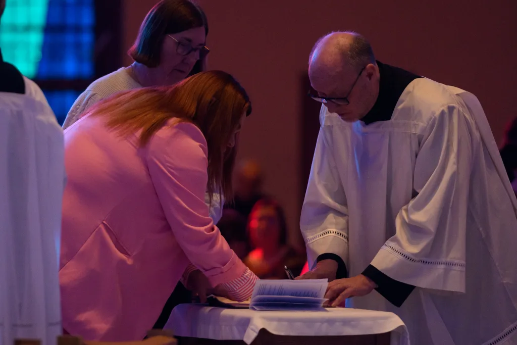 A clergyman in a white robe and a woman in a pink jacket lean over a small table in a dimly lit church as she signs a book while another woman looks on.