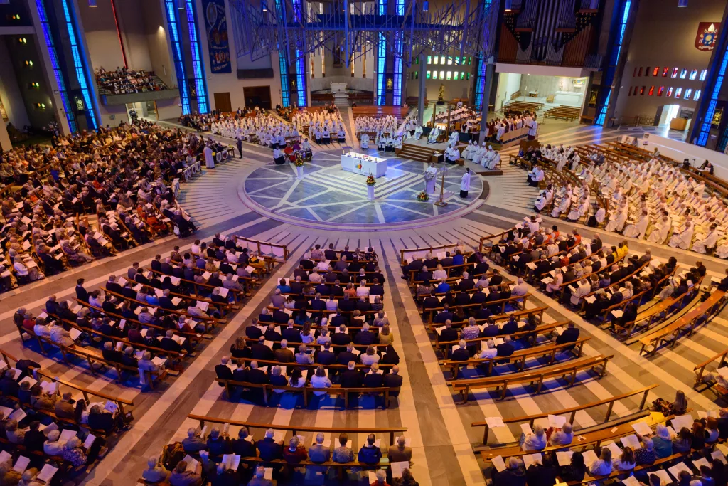 Large congregation seated in radial wooden pews inside a modern circular cathedral, gathered around a central altar where clergy in white robes officiate, framed by tall blue stained-glass windows and visible organ pipes.