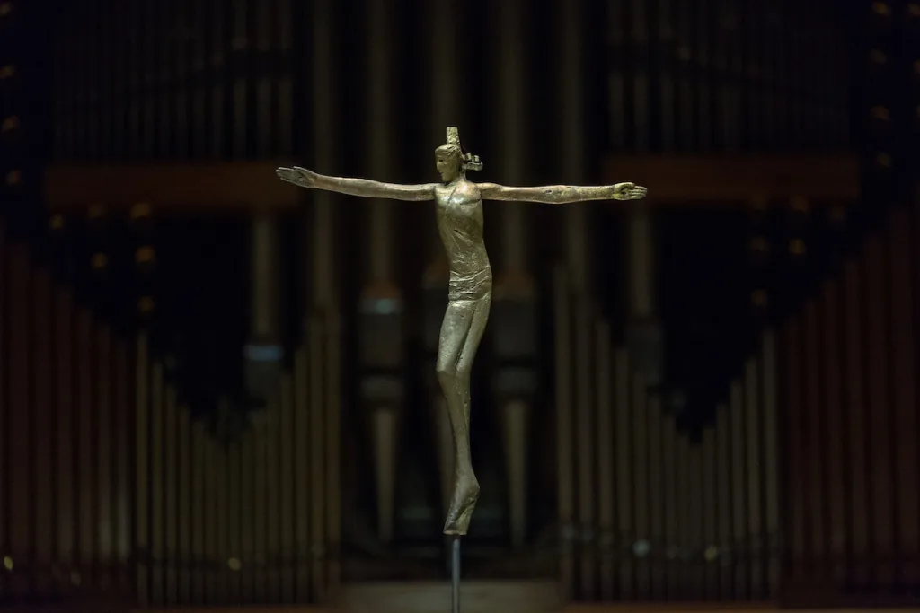 Slim bronze stylised crucifix figure with elongated body and outstretched arms mounted on a thin stand, set against a dark, blurred background of organ pipes in a church interior.