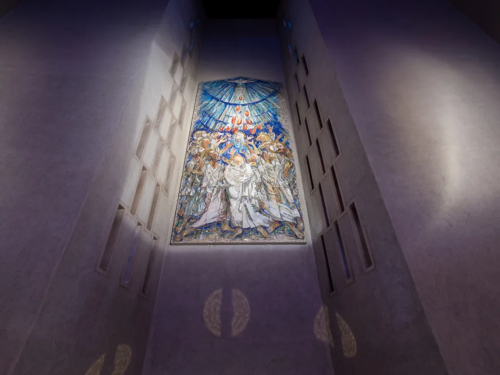 A tall mosaic mounted high in a narrow chapel alcove depicts robed figures with raised hands as red-orange tongues of flame descend from a radiant blue semicircular sky, lit from above and framed by narrow rectangular windows.