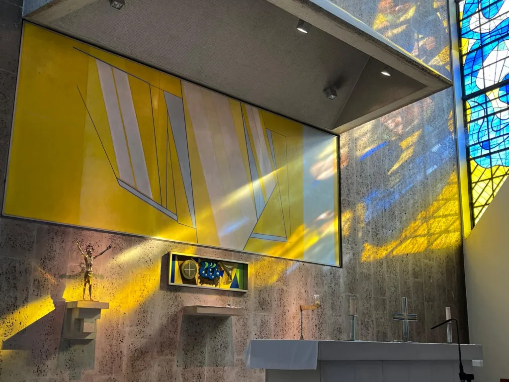Modern church interior with a white altar and glass crosses beneath a large abstract yellow mural, a bronze crucifix on a left plinth and blue‑and‑yellow stained‑glass light casting colourful reflections on the stone wall.