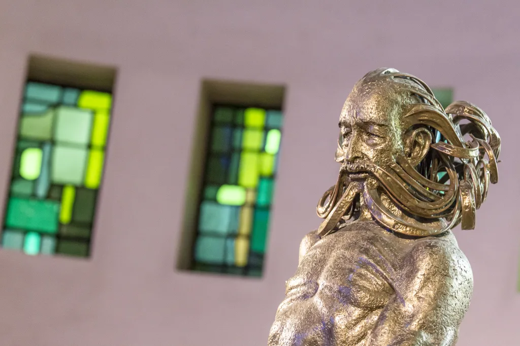 Bronze sculpture of a muscular male torso and weathered face with long, ribbon-like flowing metal beard and hair, set against blurred green-and-yellow stained-glass windows.