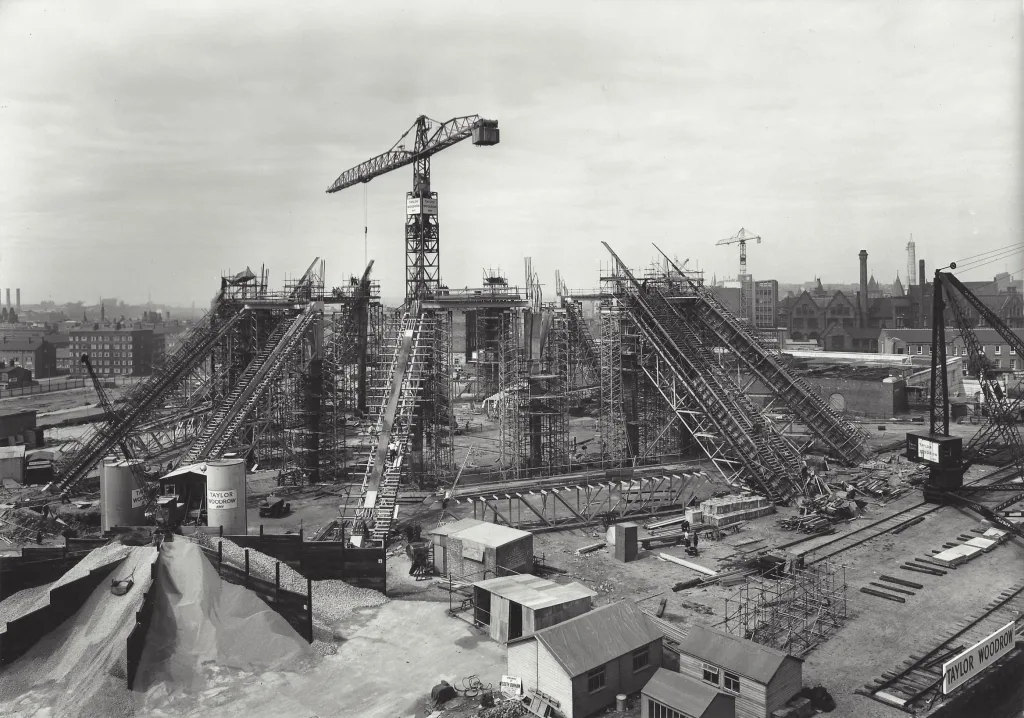 Wide black-and-white photograph of a large urban construction site with a central tower crane and several steeply inclined steel scaffold trusses forming the building framework, surrounded by temporary wooden huts, piles of gravel, rail tracks and workers, with city rooftops and a visible 'Taylor Woodrow' sign in the scene.