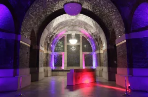 Vaulted brick-arched interior with a central chandelier, coloured purple and pink uplighting highlighting arched niches and a stained-glass window, and a curved reception desk labelled Welcome in the foreground