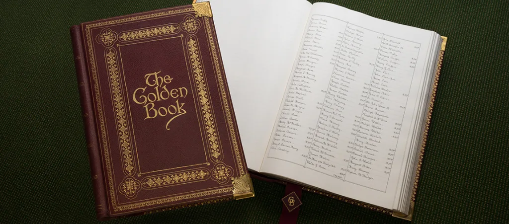 Ornate burgundy leather book titled The Golden Book with gilt decoration and corner protectors lies beside an open page showing columns of handwritten names and figures, both on a dark green textured surface.