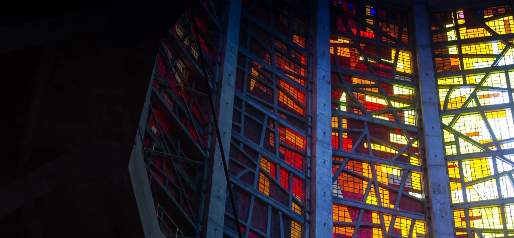 Tall stained-glass window with bold red, orange and yellow panes set in a diagonal-grid concrete and metal framework, light streaming through the abstract geometric pattern and contrasting with the dark interior.