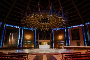 Interior of a large circular church showing a central white altar on a raised round marble platform beneath a dramatic radial metal chandelier, blue vertical stained-glass windows and organ pipes behind, with rows of wooden pews in the foreground.