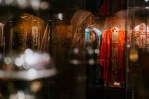 Museum display of historical liturgical robes behind glass, featuring a bright red embroidered vestment beside a multicoloured patchwork robe with ornate detailing, dim gallery lighting and soft reflections and bokeh on the glass.