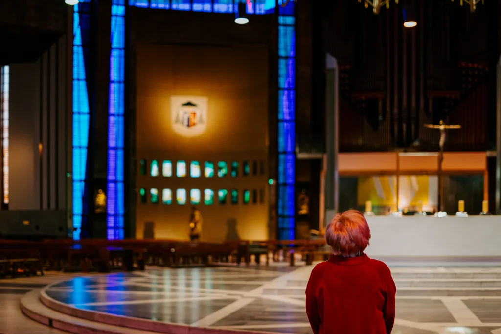 A person with short reddish hair and a red coat stands alone facing the altar inside a large church, framed by tall blue stained-glass columns, a crucifix above the sanctuary and a patterned marble floor.