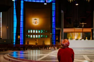 A person with short reddish hair and a red coat stands alone facing the altar inside a large church, framed by tall blue stained-glass columns, a crucifix above the sanctuary and a patterned marble floor.