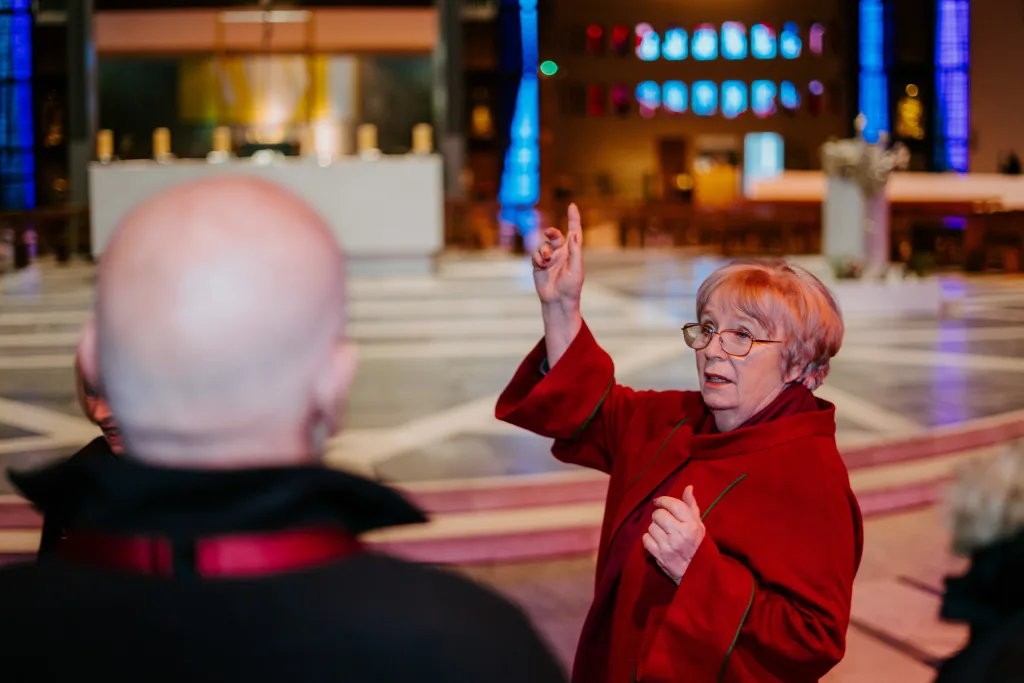 Elderly woman in a red coat and glasses gestures upward while speaking inside a church nave with an altar and stained-glass windows in the background, a blurred bald person's head in the foreground