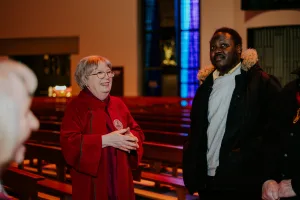 An elderly woman in a red choir robe and glasses smiles and talks with a man in a dark winter coat with a fur-trimmed hood inside a church nave with wooden pews and blue stained-glass windows behind them.