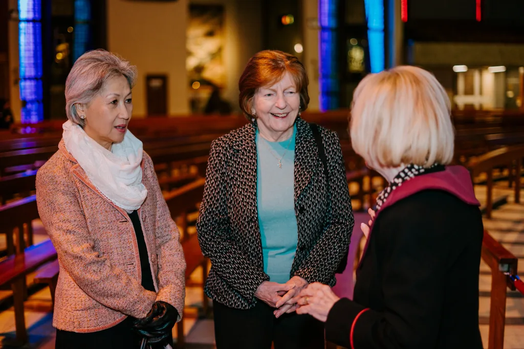 Three older women stand and chat inside a church nave with wooden pews and blue stained-glass windows; an Asian woman in a pink jacket and white scarf listens, a smiling woman in a patterned jacket and turquoise top faces the camera, and a blonde woman with a maroon-collared coat is seen from behind.