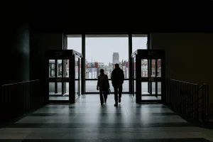 Two silhouetted people walk through a dimly lit lobby toward large glass doors that open onto a grey cityscape with a distant cathedral-like tower, framed by tiled floor and exit enclosures.