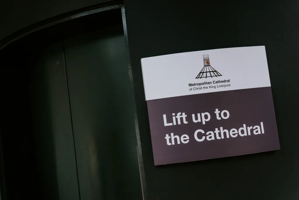 Wall-mounted sign beside dark lift doors reading "Lift up to the Cathedral" with the Metropolitan Cathedral of Christ the King Liverpool logo above the text.