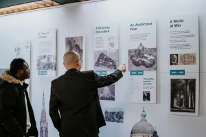 Two men stand before a museum-style timeline of large informational panels about a cathedral; a bald man in a dark suit points to a panel titled 