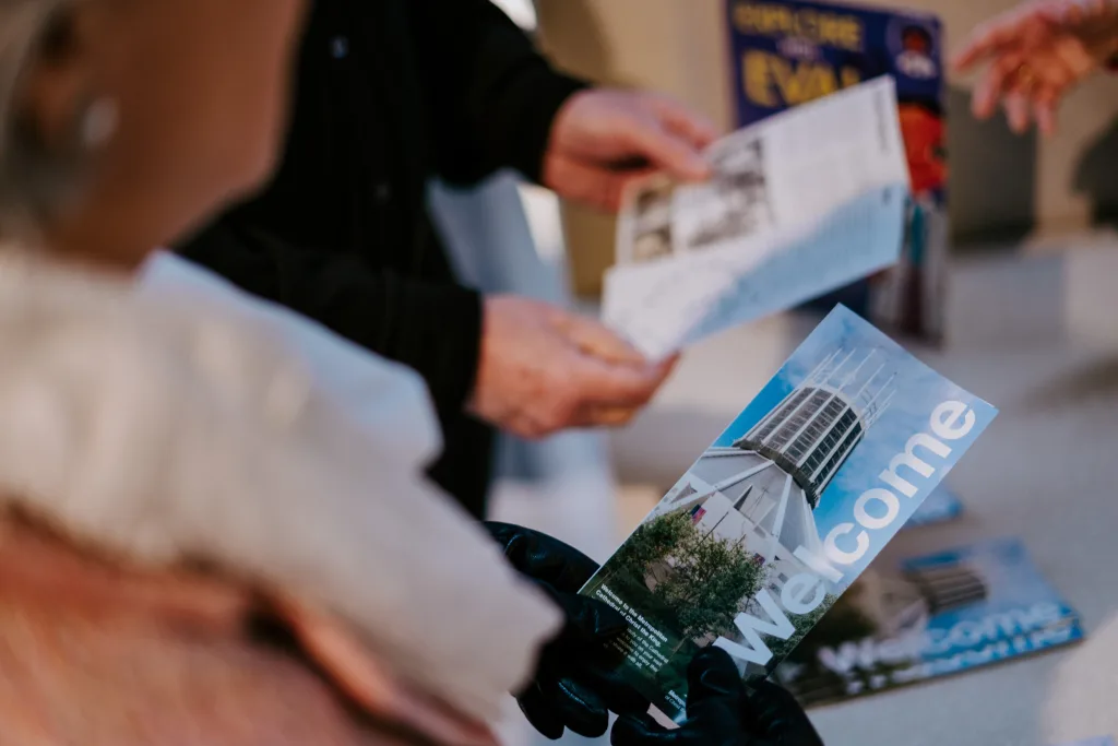 A person wearing black gloves holds a 'Welcome' brochure showing a tall modern tower, while blurred hands in the background pass out similar leaflets.