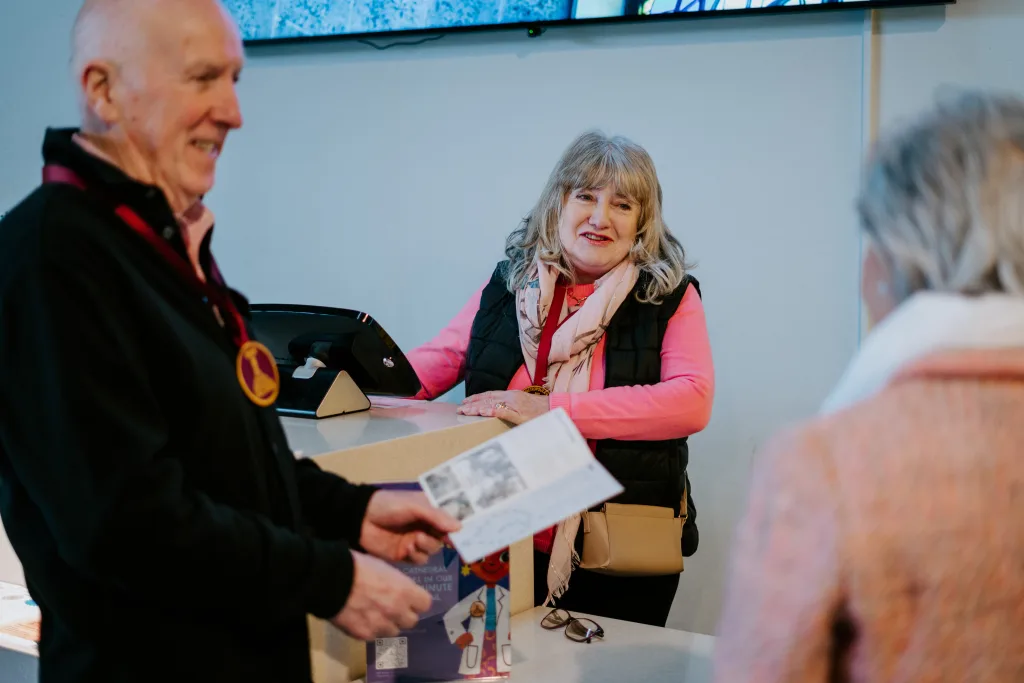 A smiling older woman in a pink jumper, patterned scarf and black quilted vest stands behind a reception desk with a beige crossbody bag and glasses on the counter as two older visitors—one blurred man holding a leaflet—talk to her.