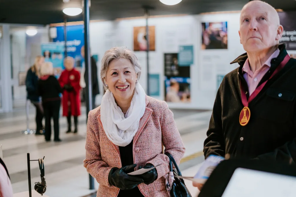 Smiling older woman in a pink tweed jacket and white scarf, wearing black gloves and holding a pamphlet, stands indoors with a man wearing a medallion to her right and blurred people and informational posters in the background.