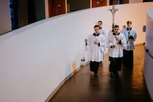 Five altar servers in white surplices and black cassocks processing down a curved church corridor, two at the front holding a swinging thurible and a small chalice while others carry tall candles and a processional cross, all with solemn expressions.