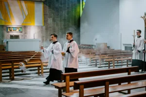 Two altar servers in white surplices process down a church nave — one swinging a smoking thurible and the other holding an incense boat — followed by servers with tall candlesticks amid rows of wooden pews, a stone altar and a stained-glass panel in the background.