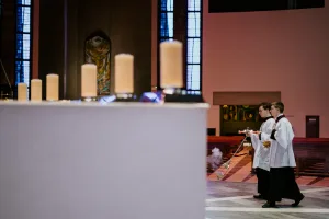 Two altar servers in white surplices and black cassocks walk down a church aisle, one swinging a thurible that emits incense, with a row of blurred candles in the foreground and tall stained-glass windows behind them.