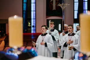 Three altar servers in white robes and red cords process through a church, one holding a censer, another a lit candle and another a small chalice, with large candles in the foreground and stained-glass windows behind them.
