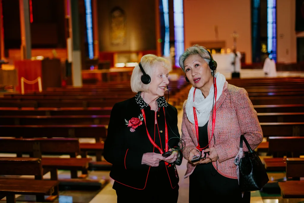 Two older women wearing audio-guide headsets and red lanyards stand and talk between wooden pews inside a church with stained-glass windows, one smiling as they hold small audio devices.