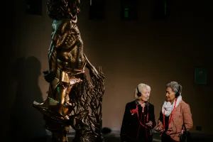 Bronze statue with intricate metalwork stands left in dim gallery light while two elderly women on the right, wearing audio-guide headphones and red lanyards, converse as they view the artwork.