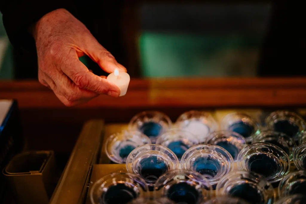A wrinkled hand holding a lit white tealight candle above a tray of clear glass votive holders filled with blue wax.