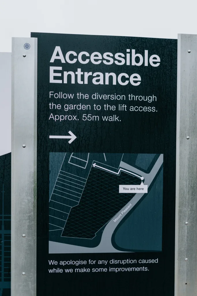 Tall black sign mounted between metal posts reading 'Accessible Entrance' with a right-pointing arrow, instructions to follow a diversion through the garden to lift access (Approx. 55m walk) and a small site map marked 'You are here'.