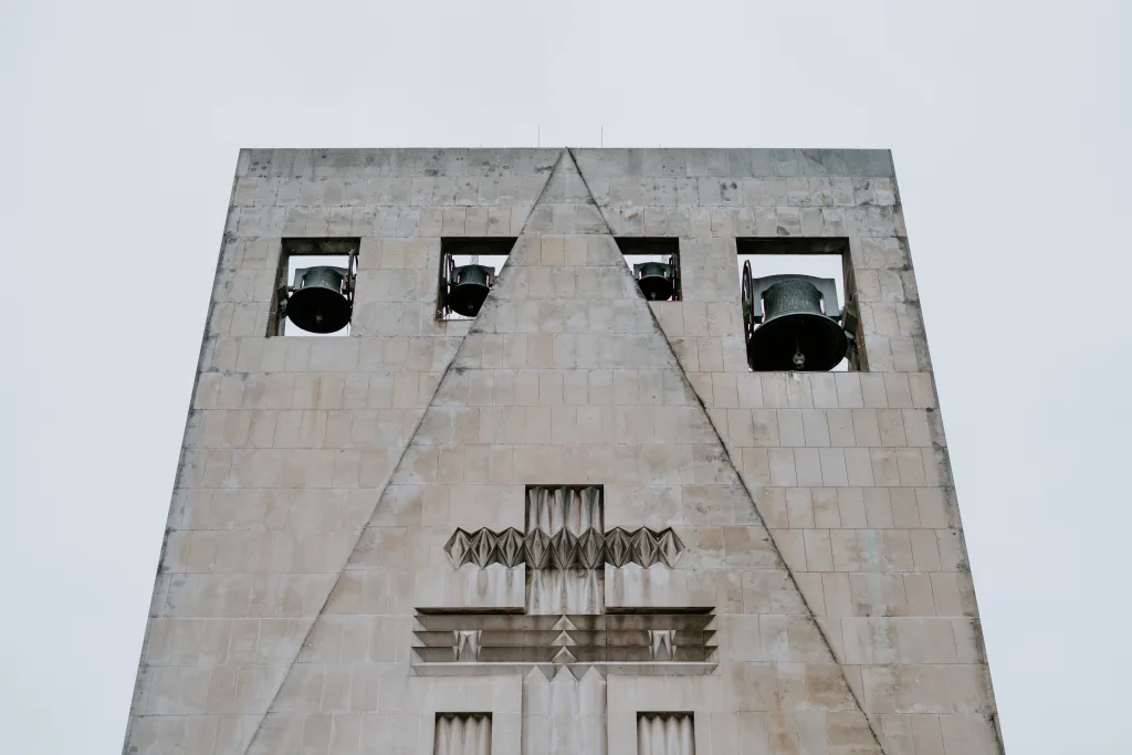 Pale stone bell tower with four square openings near the top revealing large metal bells, a central triangular groove running down the façade and angular geometric reliefs below against a pale sky.