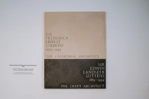 Two‑tone rectangular memorial plaque on a white wall with a beige upper panel and a dark lower panel naming two architects with their birth and death years and the inscriptions "The Cathedral Architect" (1908–1984) and "The Crypt Architect" (1869–1944), plus a small designer credit to the left.