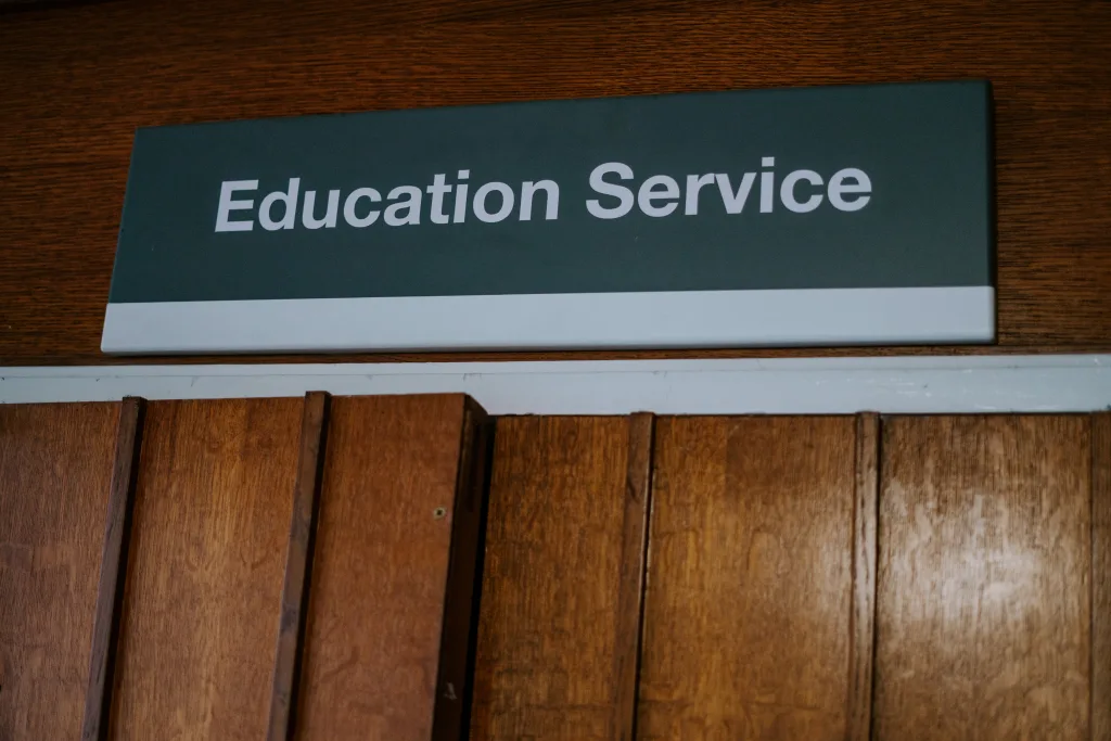 Rectangular dark green sign with white text Education Service mounted above wooden paneled doors.
