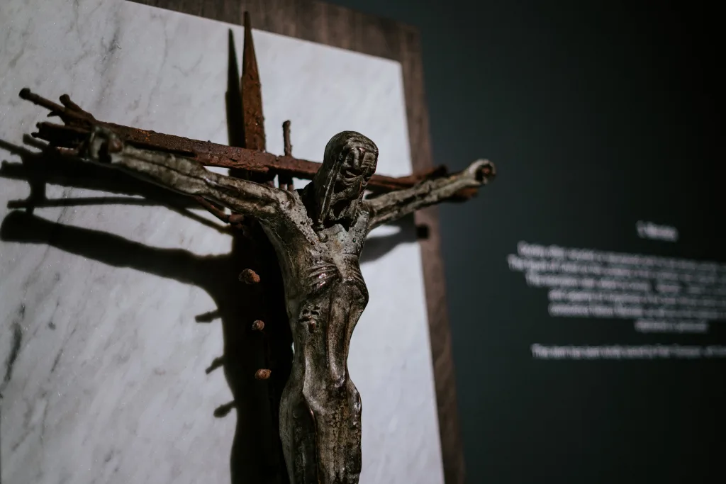 Weathered metal crucifix showing an emaciated Christ affixed to a rusted iron cross on a marble backing, casting a strong shadow against a dark gallery wall with blurred explanatory text in the background.