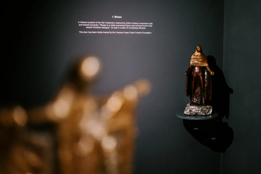Bronze and gold-toned statue of a robed, standing Moses displayed on a small wall-mounted shelf casting a shadow on a dark gallery wall, with a blurred golden figure in the foreground and a projected label titled “1. Moses” above.