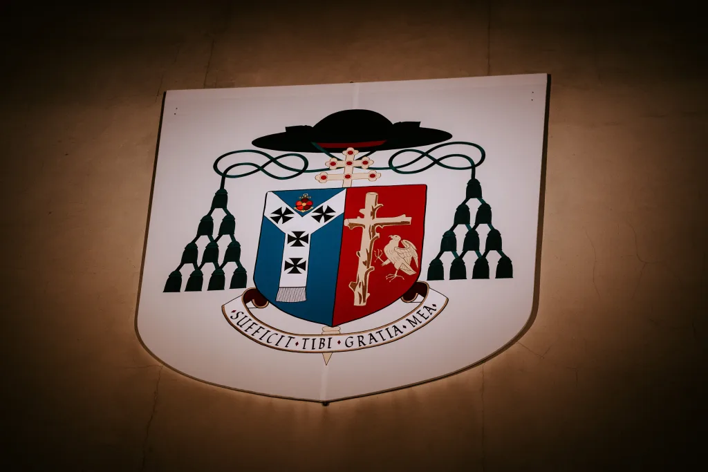 Heraldic coat of arms painted on a white shield mounted on a beige wall, showing a blue-and-red split shield with a white cross and a bird, topped by a black ecclesiastical hat with green tassels and a ribbon bearing the Latin motto SUFFECIT TIBI GRATIA MEA.