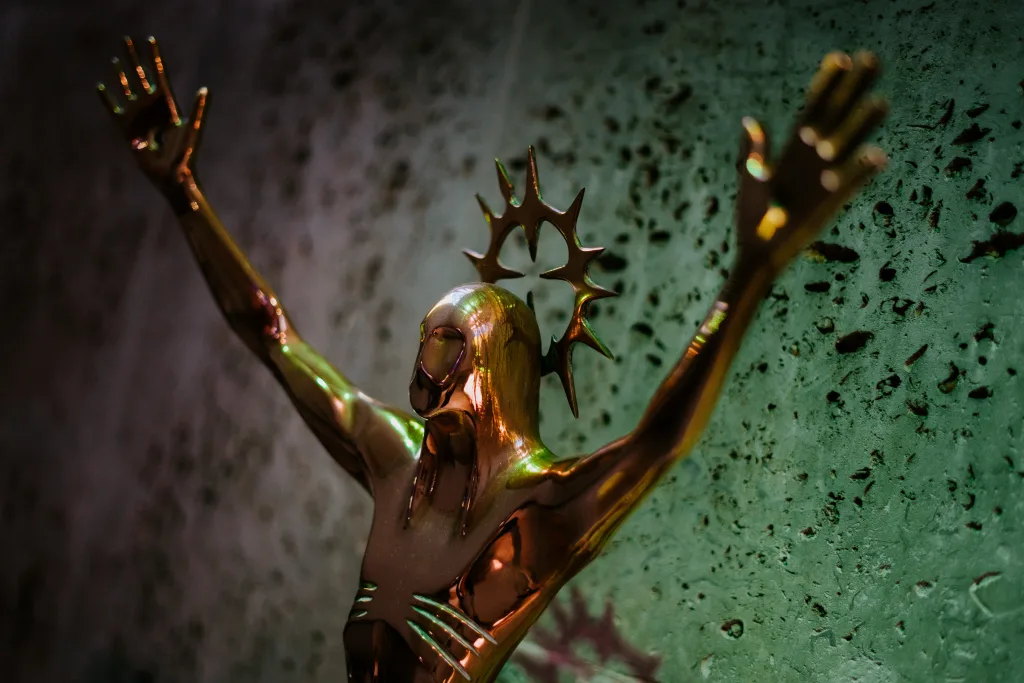 Shiny bronze statue of a stylised Christ-like figure with arms raised and a spiky halo, shown close-up against a textured green wall.