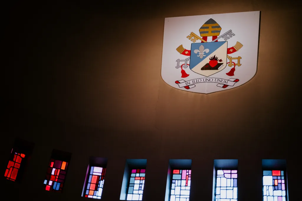 A papal coat of arms with a mitre, crossed keys and a shield showing a fleur-de-lis and a flaming heart spotlighted on a pale church wall above a row of narrow coloured stained-glass windows.