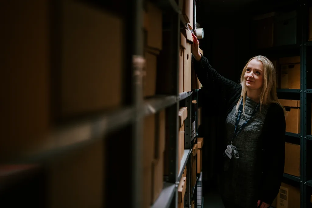A woman wearing a lanyard and dark cardigan reaches up to pull a labelled cardboard box from metal shelving in a dimly lit archive storage room filled with stacked boxes.