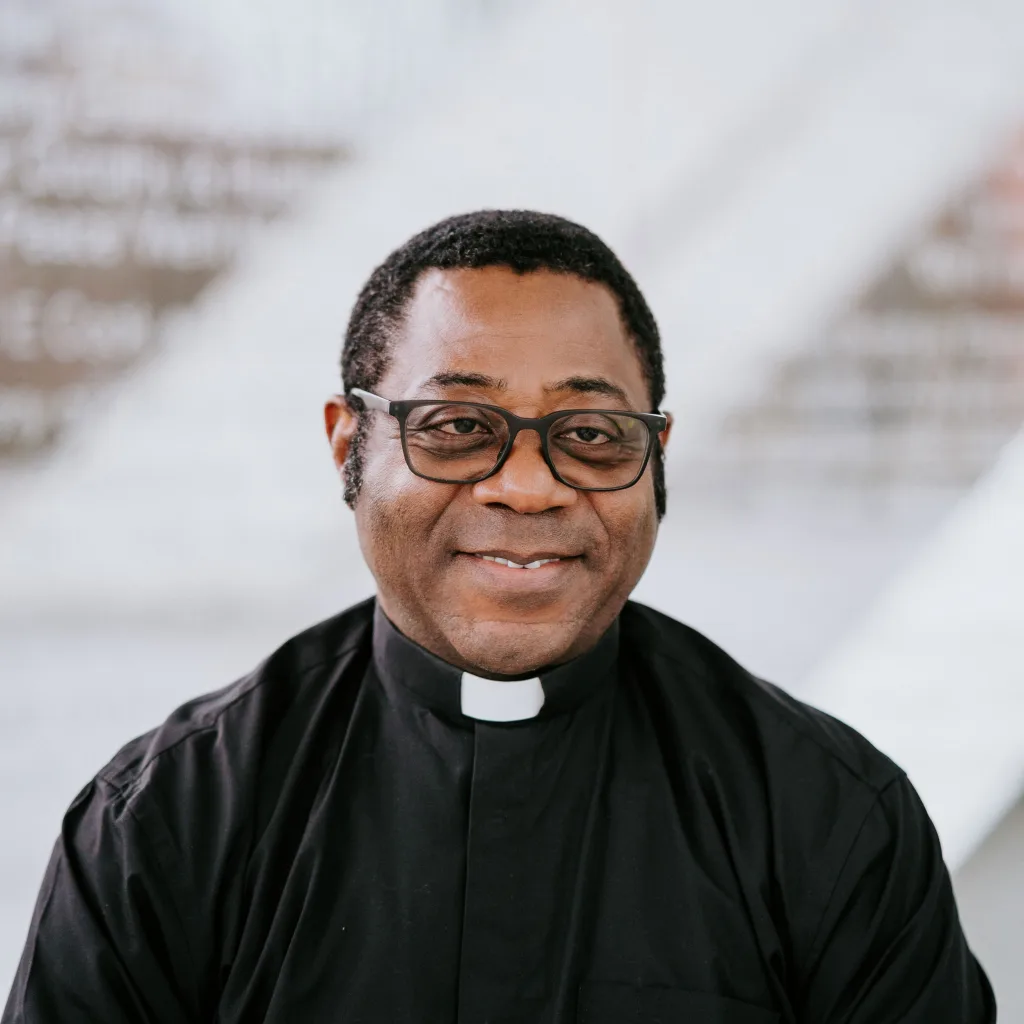Smiling man wearing rectangular glasses and a black clerical shirt with a white tab collar, photographed chest‑up against a soft, out‑of‑focus light background.