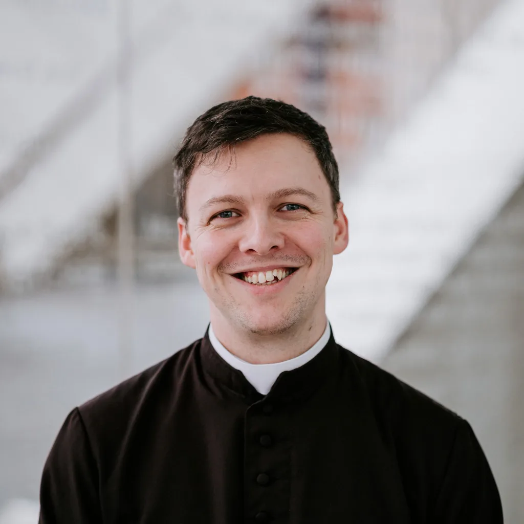 Smiling young man with short dark hair wearing a black clerical shirt and white collar, shown from the chest up against a softly blurred modern interior with diagonal lines.