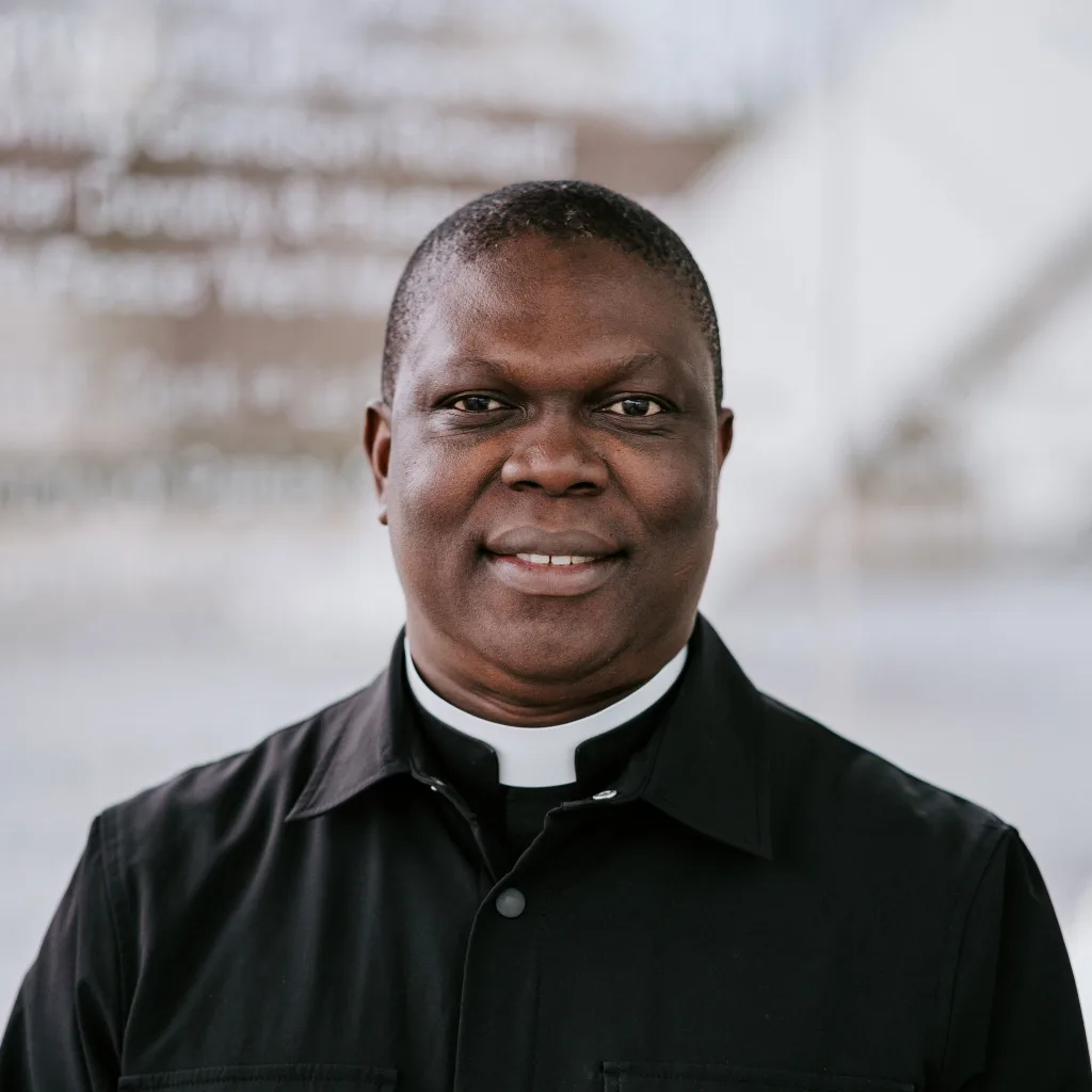Close-up portrait of a smiling Black clergyman with short hair wearing a black clerical shirt and white collar against a softly blurred light-coloured architectural background.
