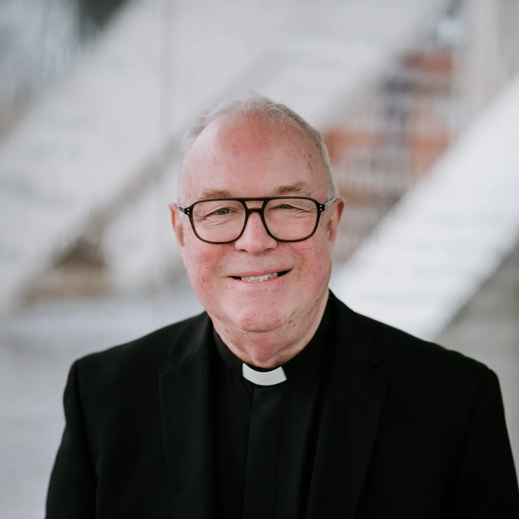 Older clergyman in a black suit and white clerical collar, smiling at the camera with short grey hair and black-framed glasses against a softly blurred staircase background.