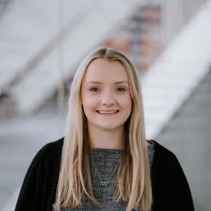Young blonde woman smiling at the camera, wearing a grey top, black cardigan and a delicate double-heart necklace, with long straight hair and a softly blurred modern staircase and architectural background.