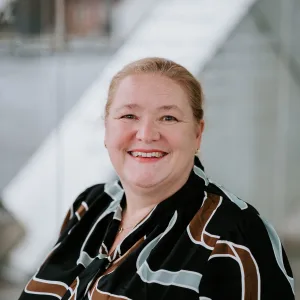 Smiling middle-aged woman with hair pulled back, wearing a black patterned blouse with brown and pale-blue geometric lines, posed against a softly lit, blurred modern architectural background.