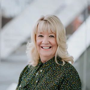 Smiling middle-aged woman with shoulder-length blonde hair and bangs wearing a dark floral blouse and a small cross pendant, photographed from the shoulders against a softly blurred light background with diagonal architectural lines.