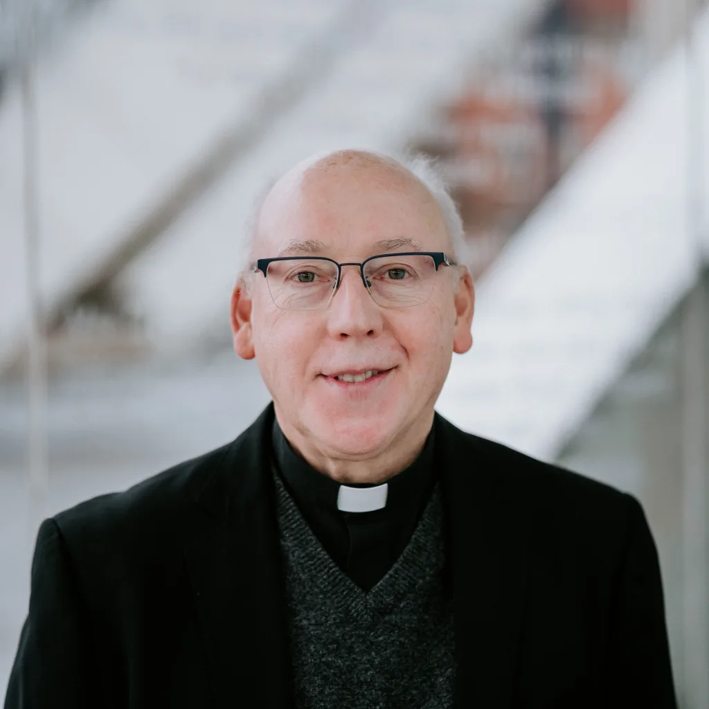 Smiling elderly man wearing glasses, a white clerical collar, dark blazer and grey sweater, framed against a softly blurred modern interior.